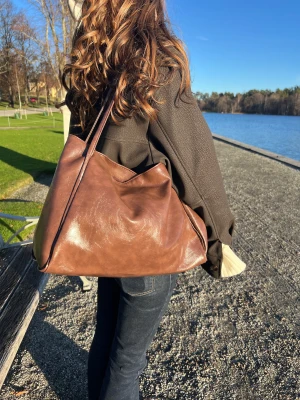 Stor brun axelväska i skinn - En stor och mjuk brun axelväska i skinn med enkel design och långa axelband. Perfekt för dig som vill ha plats för allt du behöver under dagen. Väskan har en snygg, slouchy form och passar till många olika stilar.