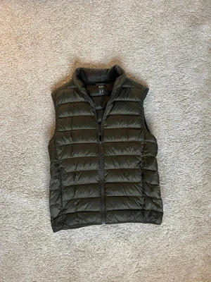 Olivgrön dunväst från Uniqlo XS - Säljer en olivgrön dunväst från Uniqlo i storlek XS. Västen är quiltad med horisontella sömmar, har hög krage och dragkedja framtill. Tillverkad i lätt syntetmaterial med dunfyllning, perfekt för lager-på-lager under kyliga dagar.
