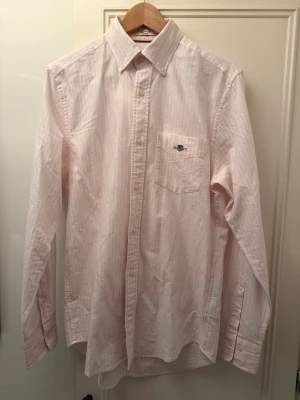 Ljusrosa randig skjorta från GANT - Säljer en ljusrosa oxfordskjorta från GANT med diskreta ränder, klassisk button down-krage och bröstficka med broderad logga. Skjortan är i regular fit och tillverkad i bomull. Perfekt för dig som gillar stilren och tidlös look.