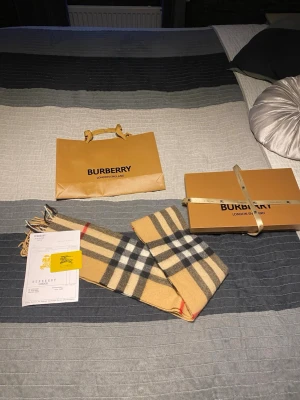 Rutig beige Burberry kashmir scarf - Exklusiv scarf från Burberry i klassiskt rutigt mönster med beige, svart, vit och röd färg. Scarfen är tillverkad i 100% kashmir. Märket och ursprung är tydligt markerat på etiketten. Perfekt för dig som preoriterar stil och elegans.
