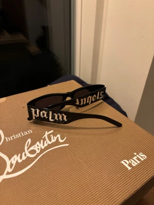 Palm Angels svarta solglasögon - Svarta solglasögon från Palm Angels med rektangulära bågar och tydlig logga i silver på sidorna. Glasögonen har en cool, urban vibe och är tillverkade i blank plast. Perfekta för att lyfta din streetwear-look.