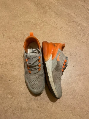 Nike Air Max 270 grå och orange - Nike Air Max 270 sneakers i grått med orange detaljer och synlig Air Max-sula. Skorna har snörning, mesh-överdel och en chunky platt sula. Perfekt för dig som gillar sportig stil och vill sticka ut med färg. Snyggt Nike-märke på sidan och häl.