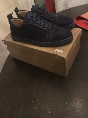 Mörkblå sneakers från Louboutin - Säljer ett par stilrena mörkblå sneakers från Louboutin med klassisk röd sula och snygg snörning. Skorna är tillverkade i mocka och har rund tå samt platt sula. Perfekt för dig som gillar exklusiva detaljer och vill sticka ut med en lyxig touch.