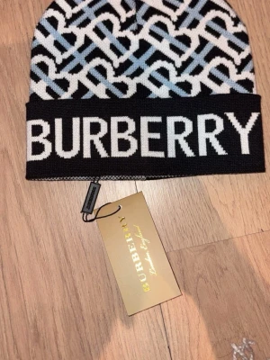 Burberry mönstrad stickad mössa - Snygg stickad mössa från Burberry med grafiskt mönster i svart, vitt och ljusblått. Stor logotyp framtill och klassisk Burberry-tag. Perfekt accessoar för att lyfta din vinterstil.
