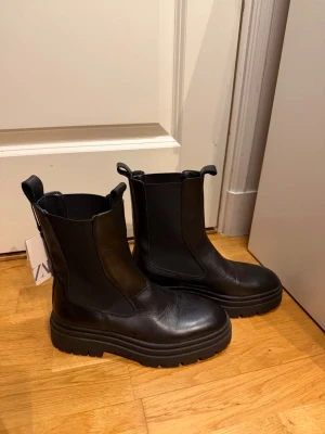 Boots från Zara - Oanvända chunky svarta chelsea boots från Zara med grov platt sula och rund tå. Tillverkade i högkvalitativt skinn med elastiska paneler på sidorna och dragflikar fram och bak. Airfit teknisk innersula i latex för extra komfort. Perfekt för dig som gillar stilrena och robusta boots. Nypris: 899kr