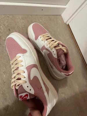 Nike Air Jordan 1 rosa och vita sneakers - Nike Air Jordan 1 sneakers i en snygg kombination av ljusrosa och vitt med beige snörning. Skorna har en klassisk låg siluett, perforerad tå och rosa yttersula. Materialet är en mix av skinn och syntet, med tydlig Nike-logga på sidan och Air-märke på plösen.