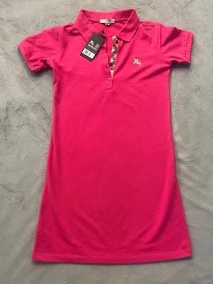 Rosa pikéktröja från Burberry - Snygg rosa pikéktröja från Burberry med klassisk krage och korta ärmar. Knappslån har Burberrys ikoniska rutiga mönster och små knappar. Klänningen har en broderad logga på bröstet och är gjord i mjuk bomull. Perfekt för en chill och stilren look.