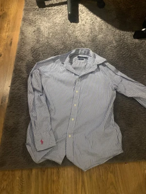 Blårandig skjorta från Polo Ralph Lauren - Snygg blårandig skjorta från Polo Ralph Lauren i custom fit. Skjortan har klassisk krage, långa ärmar och vita knappar framtill. Tillverkad i 100% bomull och har den ikoniska röda Polo-loggan broderad på manschetten. Perfekt för dig som gillar stilrena och fräscha plagg.