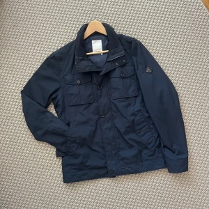Mörkblå Fieldjacket från J.Lindeberg - Söker! Hör av er om ni säljer denna jacka, köper nu! Söker i storlek S. Snygg mörkblå fieldjacket från J.Lindeberg med klassisk krage, fyra framfickor med lock och knappar samt dragkedja och knappstängning. Jackan har en stilren look och diskret logga på ärmen. Perfekt för dig som gillar enkel och clean stil.