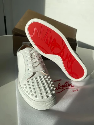 Christian Louboutin vita sneakers med nitar - Säljer ett par sjukt snygga vita sneakers från Christian Louboutin med ikonisk röd sula och coola nitar på tån. Skorna har snörning och är tillverkade i skinn, vilket ger en lyxig känsla. Perfekta för dig som vill sticka ut med en edgy och exklusiv stil.