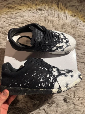 Maison Margiela skor med vitt skvätt - Maison Margiela sneakers med svart och vit färg, coolt splattermönster över hela skon. Ovandel i canvas med råa kanter och klassisk snörning. Snygg och unik design med rund tå och platt sula. Storlek 43 och inga defekter över huvud taget. Pris kan diskuteras vid snabb affär!