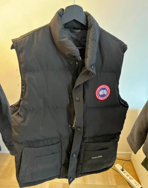 Svart dunväst från Canada Goose - Säljer en svart dunväst från Canada Goose med hög krage och klassisk logga på bröstet. Västen har stora fickor framtill, dragkedja och tryckknappar. Perfekt för kalla dagar när du vill hålla stilen enkel och clean.