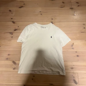 Vit t-shirt från Polo Ralph Lauren - Vit t-shirt från Polo Ralph Lauren med klassisk rund hals och korta ärmar. På bröstet sitter den ikoniska broderade polospelaren i svart. T-shirten är gjord i mjuk bomull och har en clean, stilren look som alltid funkar.