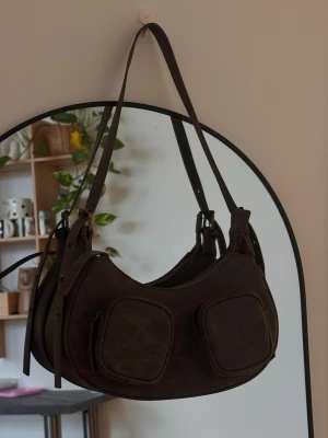 Brun axelväska i skinn med fickor - Snygg brun axelväska i skinn med två praktiska fickor framtill och justerbar rem. Väskan har rundad form och dragkedja upptill samt detaljer i antik mässing. Finns fack för kort inuti. Superfint fick!! 