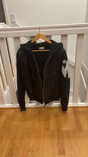 Svart Moncler pufferjacka med huva - Svart pufferjacka från Moncler med huva och dragkedja framtill. Jackan har quiltade partier på framsidan, stickade ärmar och en stor vit M-detalj på ena ärmen. Klassisk Moncler-logga på axeln och snörning i huvan. Perfekt för kyliga dagar. Den är storlek L men passar M