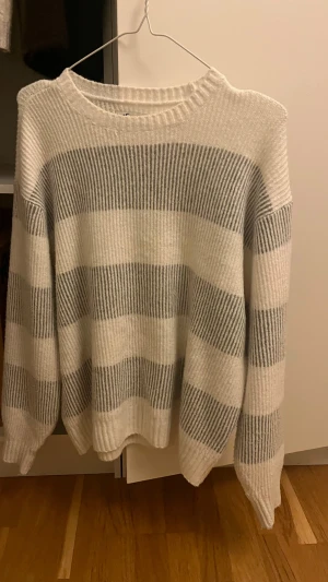 Randig stickad tröja från Hollister - Mysig stickad tröja från Hollister i storlek S. Tröjan har breda horisontella ränder i vitt och grått, rund hals och långa ärmar. Perfekt för att hålla sig varm och samtidigt se chill ut. Passar dig som gillar en relaxed vibe. Så himla skön verkligen! Lite oversized