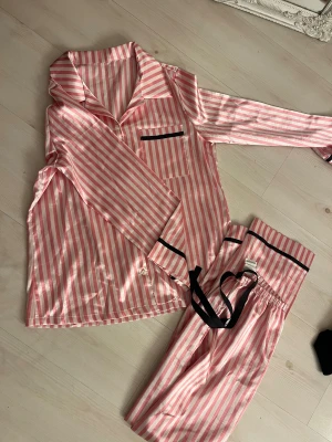 Randiga rosa pyjamasbyxor med snörning - Säljer ett par pyjamasbyxor i glansigt material med rosa och vita ränder, svart snörning i midjan och svarta detaljer. Byxorna har avslappnad passform och elastisk midja för extra komfort. Perfekt för chillkvällar hemma.