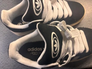 Adidas Campus svarta sneakers  - Svarta Adidas Campus sneakers med klassiska vita ränder på sidan och vit Adidas-logga på plösen. Skorna har tjocka vita snören, låg profil och gummisula i beige. Ovandelen är i mocka och insidan är vit. Perfekt för dig som gillar retrostil och streetwear.