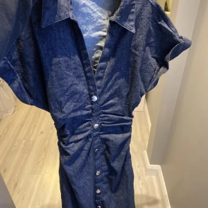 Mörkblå jeansklänning från Zara - Snygg mörkblå jeansklänning med krage och korta ärmar. Klänningen har knappar hela vägen framtill och cool rynkning vid midjan som ger en schysst siluett. Perfekt för dig som gillar denim och vill ha en unik look.
