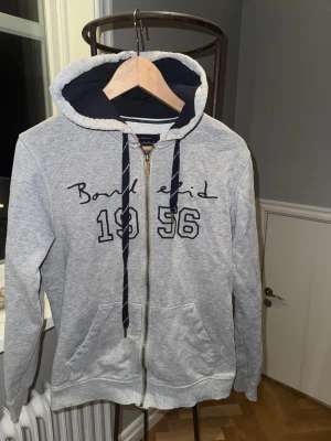 Grå hoodie från Bondelid med dragkedja - Snygg grå hoodie från Bondelid med mörkblå detaljer och broderad text samt siffrorna 1956 på bröstet. Tröjan har huva med matchande snören, två fickor fram och en rejäl dragkedja i metall. Perfekt för chill dagar och sköna promenader.