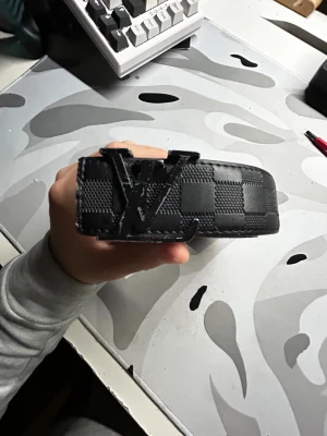 Svart rutigt L v bälte - Svart bälte från L V med klassiskt rutigt mönster och stor logga i metall på spännet. Bältet är tillverkat i skinn och har en stilren, modern look som passar perfekt till jeans eller chinos. Märkt med 'Made in France' på insidan.