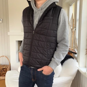 Svart quiltad dunväst John Henric - Svart quiltad dunväst från John Henric med dragkedja framtill och diskret rutmönster. Västen har en klassisk passform och är perfekt att slänga över en hoodie. Lätt och smidig, passar till många olika outfits.