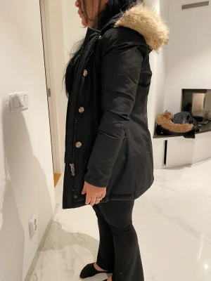 Svart parkajacka med fuskpäls - Svart parkajacka med huva och fluffig beige fuskpäls runt kanten. Jackan har långa ärmar, dragkedja och knappar framtill. Snygg, stilren och perfekt för kyliga dagar. Passformen är normal och jackan går ner över höften.