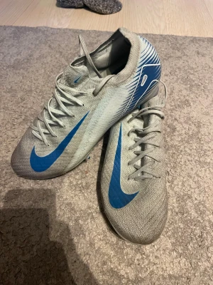 Nike Air Zoom fotbollsskor grå/blå - Säljer ett par Nike Air Zoom fotbollsskor i grått med blå detaljer och snörning. Skorna har en smal och sportig siluett med rund tå och platt sula med dobbar för extra grepp på planen. Materialet är syntet och mesh för lätthet och ventilation. Snyggt mönster på hälen och yttersulan.