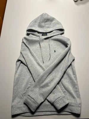 Grå hoodie från Polo Ralph Lauren - Snygg grå hoodie från Polo Ralph Lauren med klassisk broderad logga i mörkblått på bröstet. Tröjan har huva med snörning, ribbade muddar och en mjuk insida. Perfekt för chill dagar och enkel att matcha med jeans eller joggers.