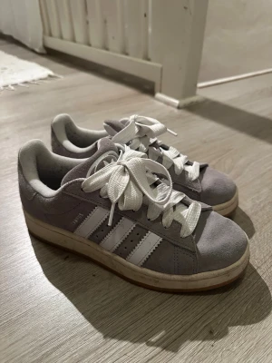 Grå Adidas Campus sneakers strl 39 - Säljer ett par grå Adidas Campus sneakers med klassiska vita ränder på sidorna och vit sula. Skorna har rund tå, platt sula och är tillverkade i mjuk mocka med vita snören. Perfekt för dig som gillar retrostil och streetwear.
