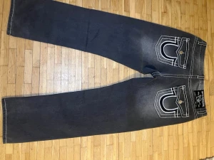 True Religion svarta jeans med kontrastsöm - Svarta jeans från True Religion med tydliga vita kontrastsömmar och ikoniska fickdetaljer bak. Klassisk femficksmodell med metallknappar och logotyp på bakfickorna. Jeansen har rak passform och är tillverkade i slitstarkt denimtyg.