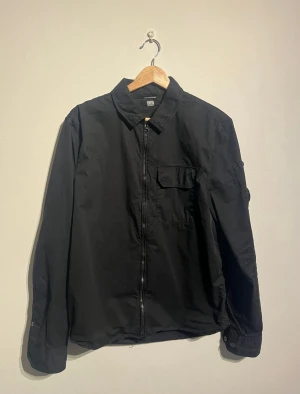 C.P Company Svart Overshirt - Svart overshirt från C.P. Company med klassisk krage och dragkedja framtill. Långärmad modell med ikonisk linsdetalj på vänster ärm. Tillverkad i bomull för en skön och stilren känsla.   Skick: Mycket gott skick, sparsamt använd.  Storlek: M men sitter som en S  Kolla gärna in mina andra annonser.   Skriv vid frågor eller ifall du vill ha fler bilder.