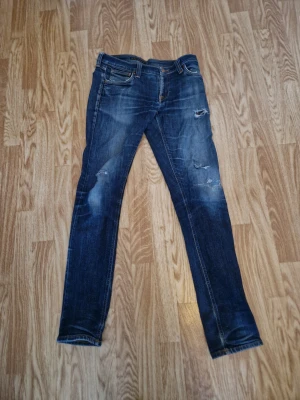 Blå skinny jeans från Nudie Jeans - Snygga blå jeans från Nudie Jeans med slitna detaljer och skinny passform. Klassisk femficksmodell med orange sömmar och Nudie-logga på insidan. Jeansen har en mörk tvätt med ljusare partier och är tillverkade i jeansmaterial. Modellen på byxorna är tight long john. Tyvärr håll inne i fickan på vänster sida som går att laga hos skräddare. 