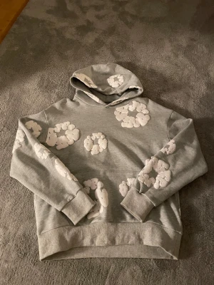 Grå Denim Tears hoodie - Säljer en grå hoodie från Denim Tears med unika vita mönster som ser ut som moln eller blommor. Tröjan har huva, långa ärmar och ribbade muddar. Materialet är mjukt och känns som bomull, perfekt för chill dagar. Hoodien har en loose passform och cool streetwear-vibe.