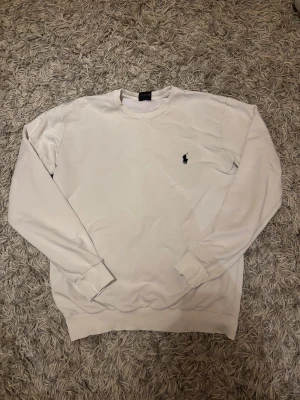 Vit sweatshirt från Polo Ralph Lauren - Vit sweatshirt från Polo Ralph Lauren med klassisk rund hals och svart broderad logga på bröstet. Tröjan har ribbade muddar vid ärmslut och nederkant, och är tillverkad i mjuk bomull. Perfekt för chill dagar och enkel att matcha med jeans eller joggers.