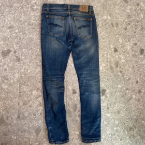 Blå Nudie jeans - Säljer ett par blå jeans från Nudie Jeans med slitningar och snygga detaljer på . orangea sömmar och Nudie-logga på bakfickan. Passar dig som gillar en smal siluett och coola slitningar.