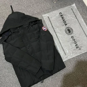Svart macmillan jacka från Canada Goose - Svart dunjacka från Canada Goose med huva och klassisk logga på ärmen. Jackan har en quiltad design, långa ärmar och knäppning framtill. Perfekt för kalla dagar och har en clean, stilren look som funkar till det mesta.