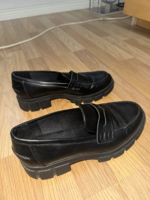 Chunky svarta loafers i skinn - Säljer ett par chunky svarta loafers med grov platt sula och rund tå. Skorna är tillverkade i skinn och har klassisk pennyloafers-design med stilren söm och detalj framtill. Köpta för cirka 500kr och aldrig använda 