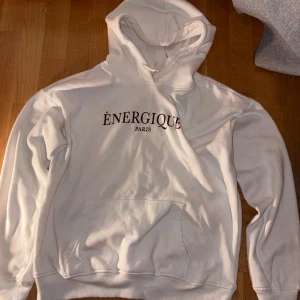 Vit hoodie Énergique Paris - Vit hoodie från Énergique Paris med tryckt logga på bröstet. Tröjan har huva med snörning och en stor magficka framtill. Mjuk och skön bomullskänsla, perfekt för chill dagar. Klassisk passform och ribbade muddar vid ärmslut och nederkant.