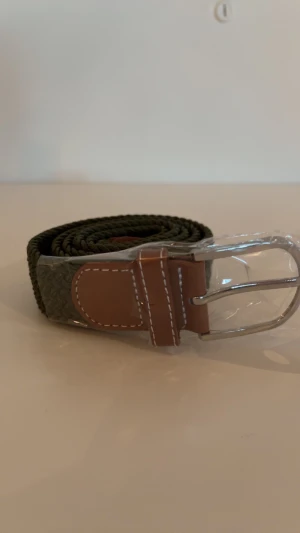 Olivgrönt flätat bälte  - Snyggt flätat bälte i mörkgrön färg med bruna skinn-detaljer och silvrigt spänne. Bältet har en grov, vävd struktur som ger en cool och modern känsla. Perfekt för att lyfta din outfit med en touch av stil. Passar till jeans eller chinos.