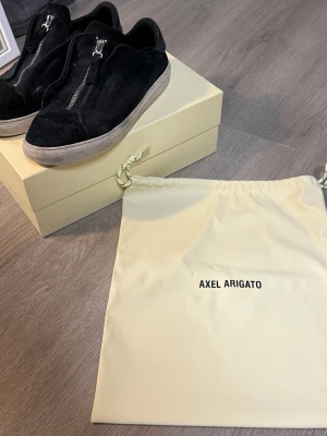Svarta sneakers Axel Arigato mocka - Svarta sneakers från Axel Arigato i mjuk mocka med dragkedja framtill istället för klassisk snörning. Skorna har en vit platt sula och minimalistisk design, perfekt för dig som gillar stilrena detaljer och streetwear-vibe.
