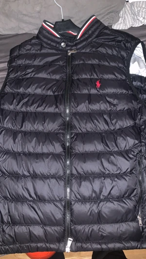 Svart Ralph Lauren Väst - Svart dunväst från Polo Ralph Lauren i storlek L med röd logga broderad på bröstet. Västen har quiltad design, dragkedja framtill och ståkrage med röd och vit randig detalj. Perfekt för lager-på-lager och snygg till streetwear. Har använts 2 gånger men den är i toppen skick 👍 Nypris: 3369kr 