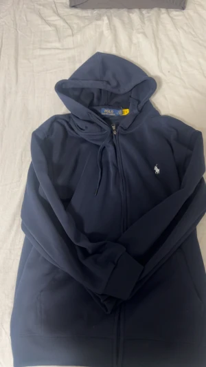 Mörkblå hoodie från Polo Ralph Lauren - Snygg mörkblå hoodie från Polo Ralph Lauren med klassisk logga broderad i vitt på bröstet. Tröjan har huva med snörning, dragkedja framtill och ribbade muddar. Perfekt för chill dagar och enkel att matcha med jeans eller joggers.