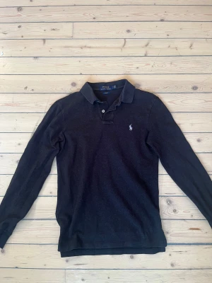 Svart långärmad pikétröja Polo Ralph Lauren - Svart långärmad pikétröja från Polo Ralph Lauren i slim fit. Klassisk krage med två knappar och den ikoniska broderade loggan på bröstet. Tröjan är tillverkad i mjuk bomull och har en stilren look som funkar till många olika outfits. Mycket bra skick