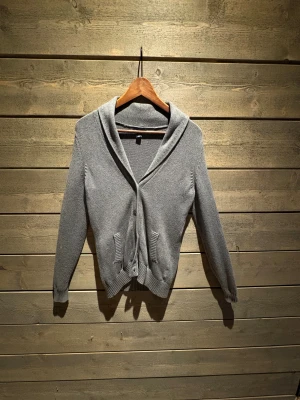 Tommy Hilfiger cardigan - Grå stickad kofta med bred krage och två framfickor från Tommy Hilfiger . Stängs med knappar framtill och har ribbade muddar vid ärmslut och nederkant. Perfekt för lager-på-lager och chill stil. Långärmad och mjuk känsla. 