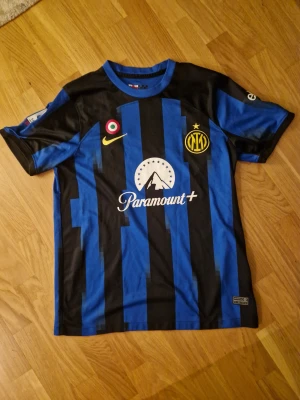 Inter Milan matchtröja Nike blå/svart - Inter Milan fotbollströja från Nike med klassiska blå och svarta vertikala ränder, gul Nike-logga och klubbmärke med stjärna. Paramount+ sponsortryck på bröstet. Rund hals och korta ärmar. Tillverkad i lätt polyester som andas.