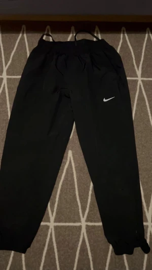 Svarta mjukisbyxor från Nike - Svarta mjukisbyxor från Nike med klassisk vit swoosh-logga på benet. Byxorna har elastisk midja med snörning och är tillverkade i mjukt syntetmaterial. Passformen är loose och benen är raka med resår vid benslut. Perfekta för chill eller träning.