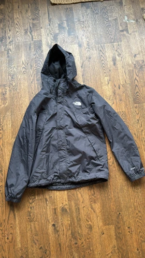 Gortex The North Face windbreaker - Svart gortex windbreaker från The North Face i storlek M. Jackan har huva med snörning, dragkedja framtill och elastiska muddar vid ärmslut. Materialet är lätt och syntetiskt, perfekt för blåsiga dagar. Logga på bröstet och ryggen ger en clean och sportig vibe.