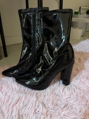 Svarta lackstövletter med klack från NLY Shoes - Säljer ett par svarta stövletter i glansigt lackmaterial från NLY Shoes. Skorna har spetsig tå, hög blockklack och dragkedja på sidan för enkel påtagning. Perfekt för dig som vill sticka ut med en edgy look.