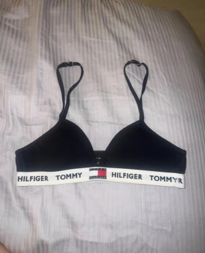 Svart bh  från Tommy Hilfiger - Svart bh från Tommy Hilfiger med justerbara axelband och vit elastisk kant med logotypen tryckt i svart. Snygg och enkel design med sportig känsla, perfekt för att styla med jeans eller shorts. Materialet är mjukt och stretchigt för skön passform. 12-13 år 